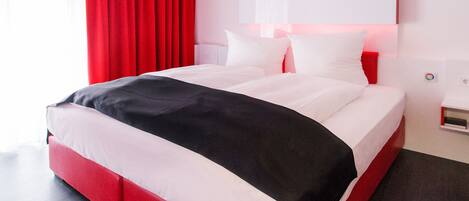 A modern hotel room with a large bed, red curtains, and a white headboard.