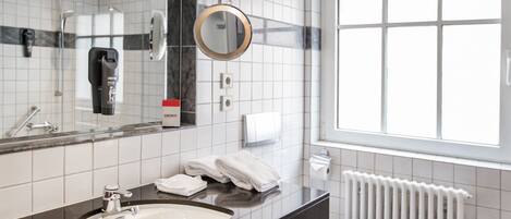 Un baño moderno con encimera negra, lavabo blanco y un espejo grande. Hay una ventana que proporciona luz natural y un radiador blanco.
