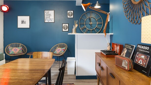 A room with a blue wall, a wooden dining table, two wicker chairs, a white cabinet, a clock, and a lamp. - O'Lodge By Arvor Hotel (Dinan)