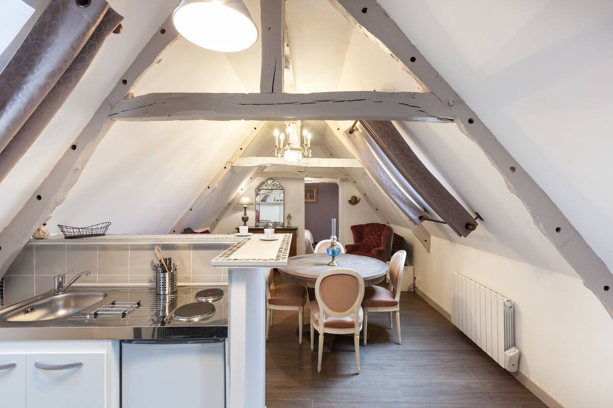 A cozy attic dining area with a round table and chairs, a chandelier, and a kitchen with a sink and utensils.