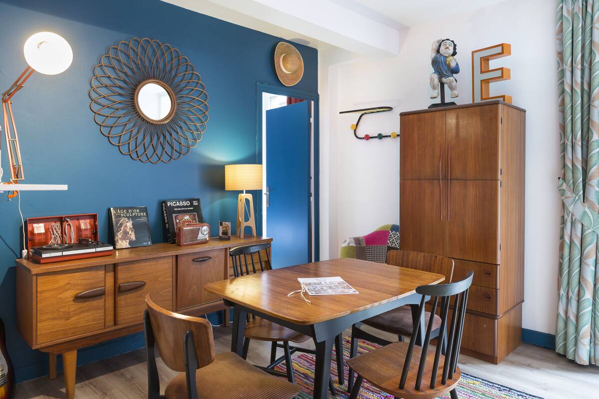 A dining area with a wooden table and chairs, a cabinet with decorative items, and a blue wall with a mirror and wall art.