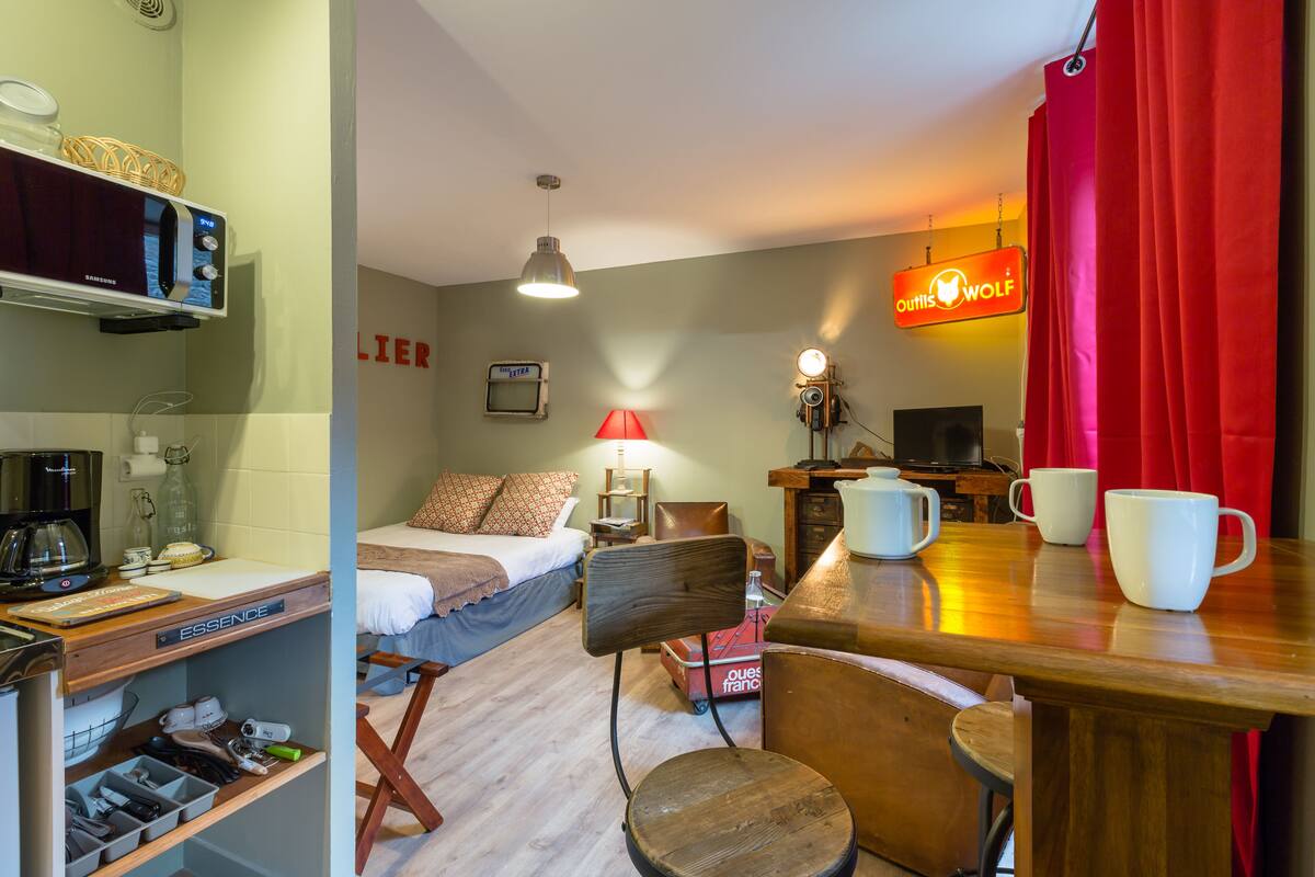 A hotel room with a bed, a desk with a computer, a chair, a table with mugs, and a red curtain.