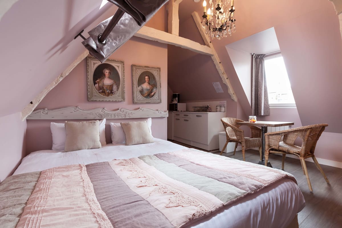 A bedroom with a large bed, a desk, and two chairs. There are two framed pictures on the wall and a chandelier.