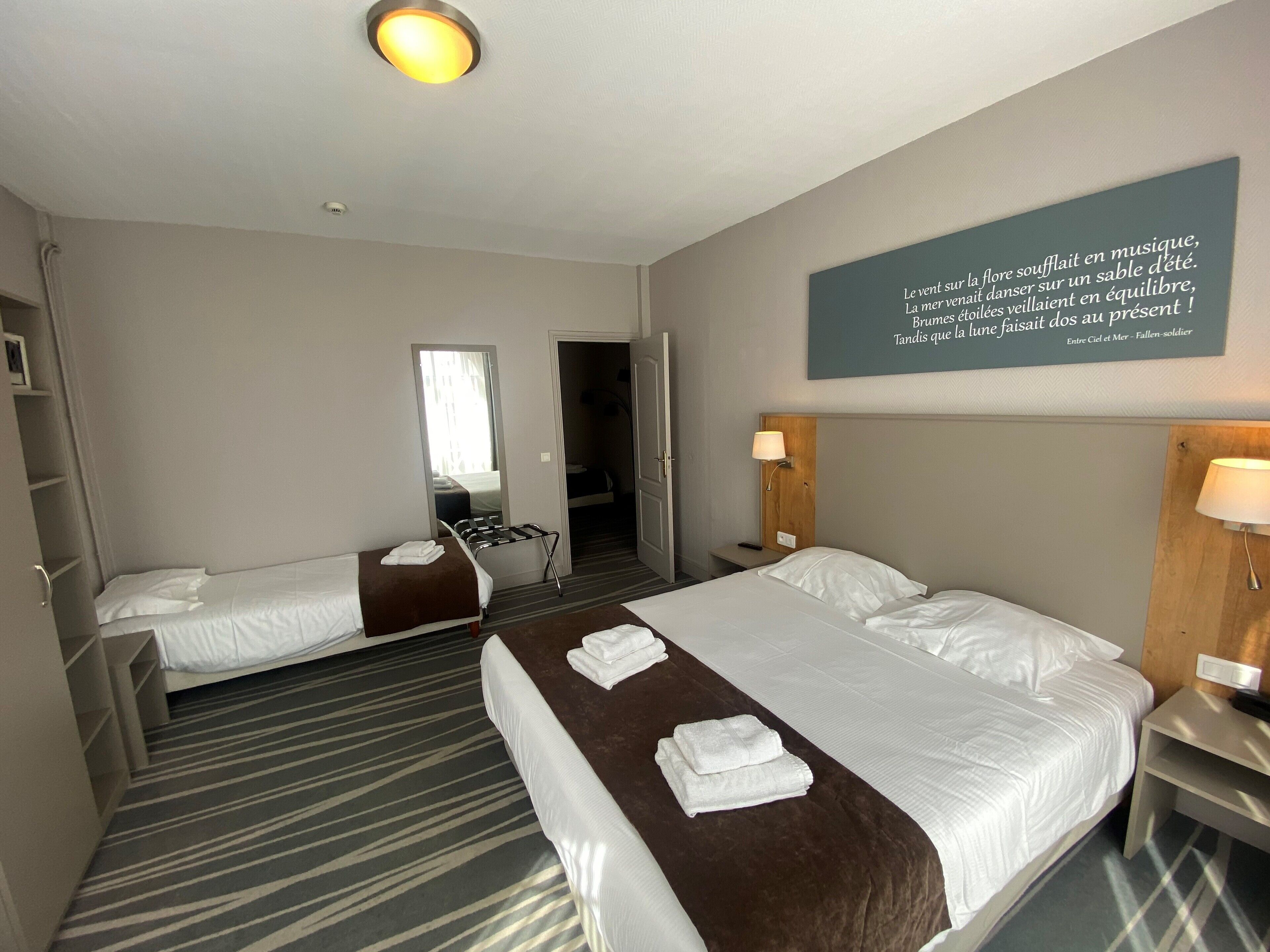 A hotel room with two beds, a large mirror, a bedside table with a lamp, and a motivational quote on the wall.