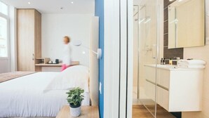 A modern hotel room with a bed, bedside table with a plant, a desk with a chair, a large mirror, and a bathroom with a shower and sink. - Hôtel Villa Des 2 Caps (Wimereux)