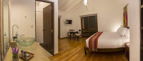 A bedroom with a glass-bottomed sink, wooden flooring, a bed with a white comforter, and a television mounted on the wall.
