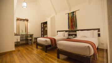 A hotel room with two beds, a wooden floor, a bathroom with a mirror and sink, and a colorful tapestry on the wall.