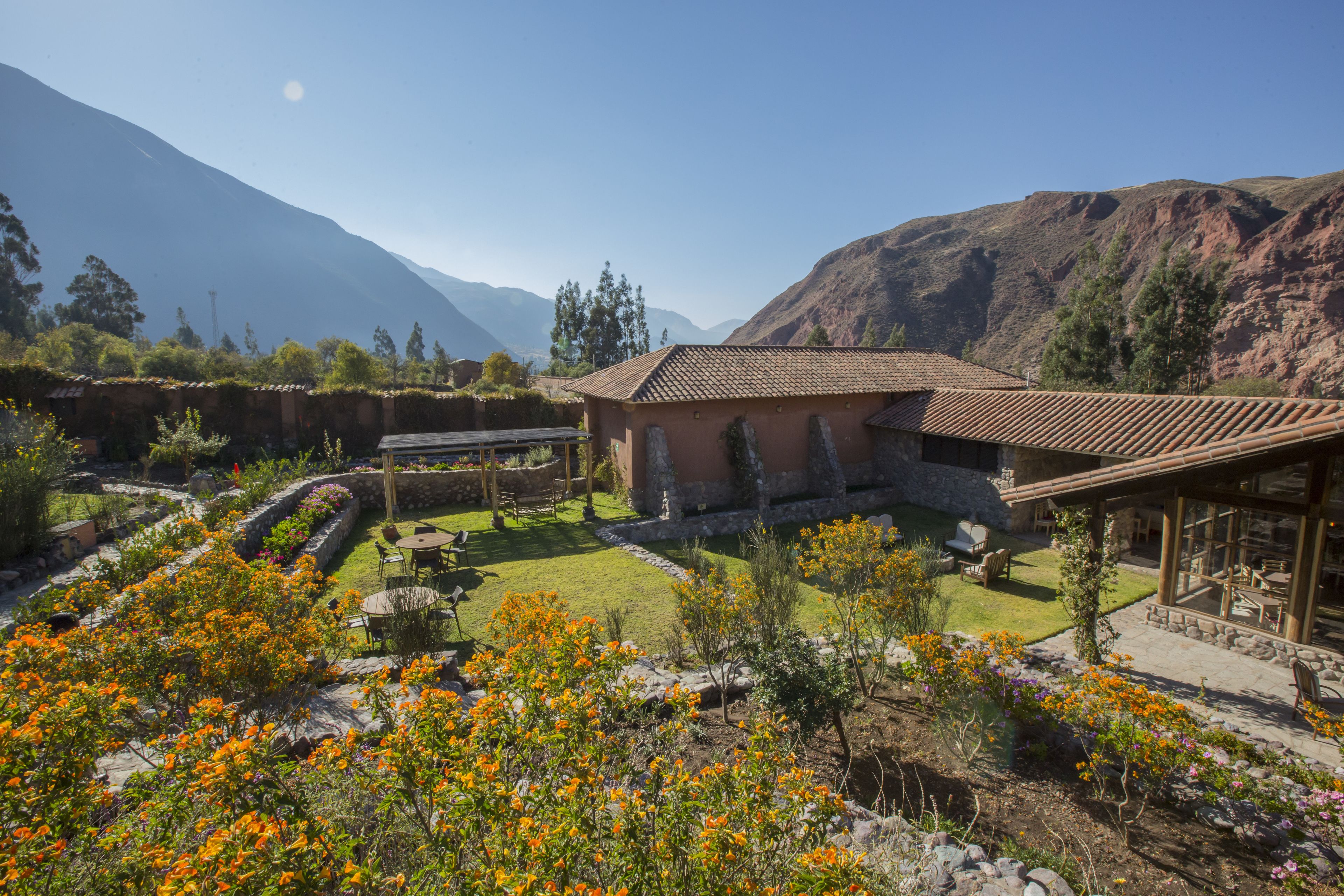 A rustic house with a tiled roof, surrounded by a well-maintained garden with flowering plants, a stone wall, and a view of mountains in the background.