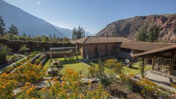 A rustic house with a tiled roof, surrounded by a well-maintained garden with flowering plants, a stone wall, and a view of mountains in the background.