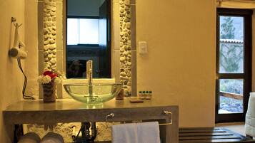 Un baño con pared de piedra, un lavabo de vidrio y un espejo grande. Hay toallas en la encimera y una ventana con vistas a árboles.
