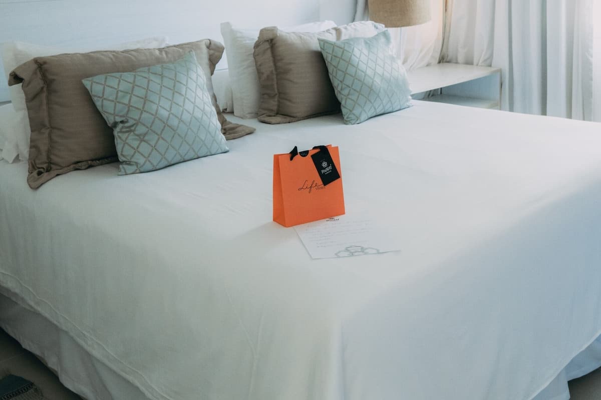 A hotel room with a bed, pillows, a nightstand, a lamp, and an air conditioning unit.