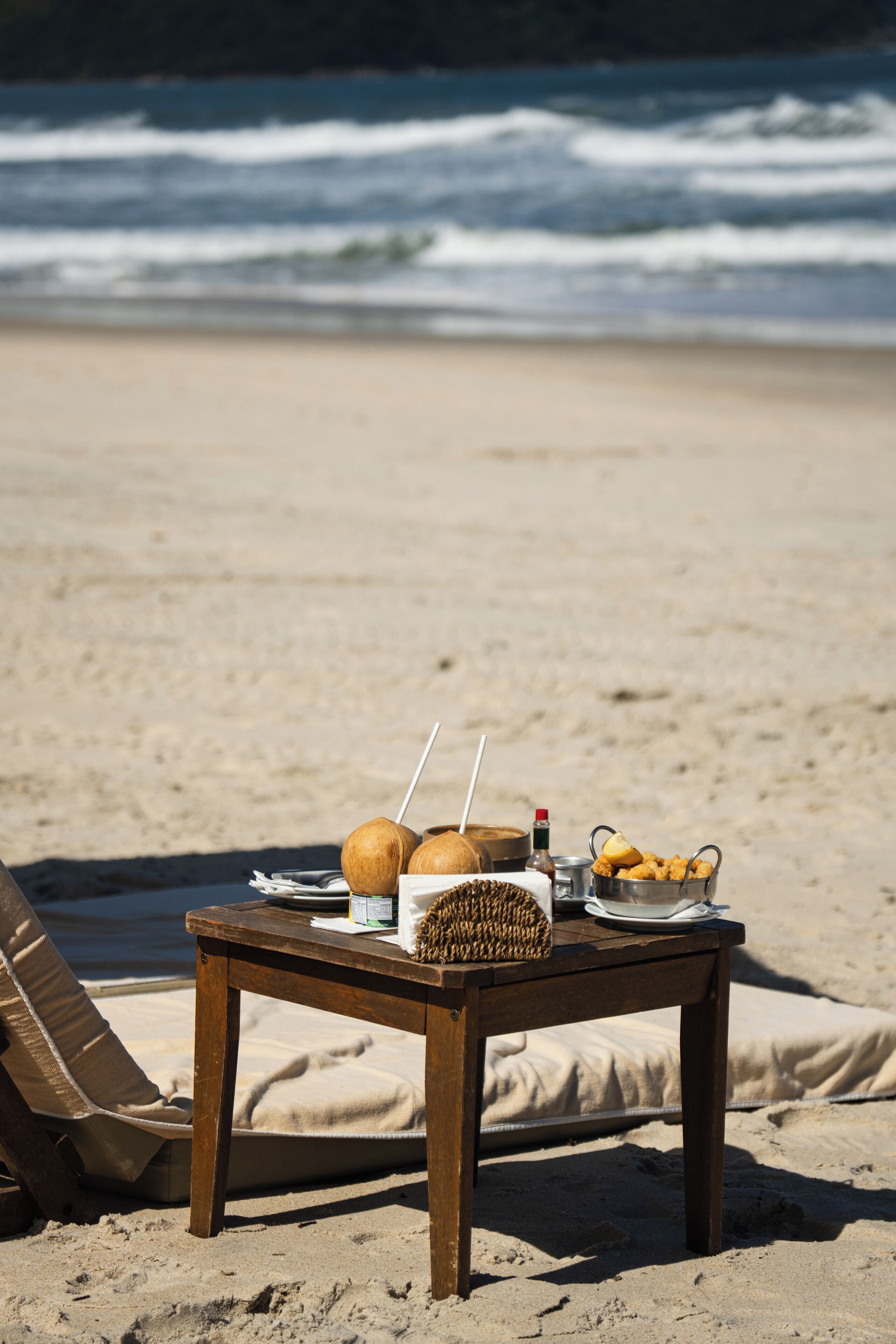 Una mesa de madera con una cesta, cocos, una bebida y aperitivos en la playa.