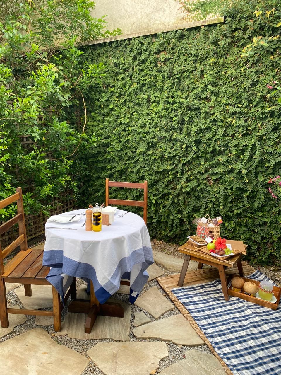 An outdoor patio with a round table covered by a white tablecloth with blue edges, surrounded by wooden chairs. A small wooden table with fruits and a basket is placed against a wall covered in green ivy.