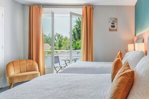 A hotel room with a bed, a chair, a balcony with a table and chairs, and a view of greenery. - Hôtel l'Amandier (Morlaas)