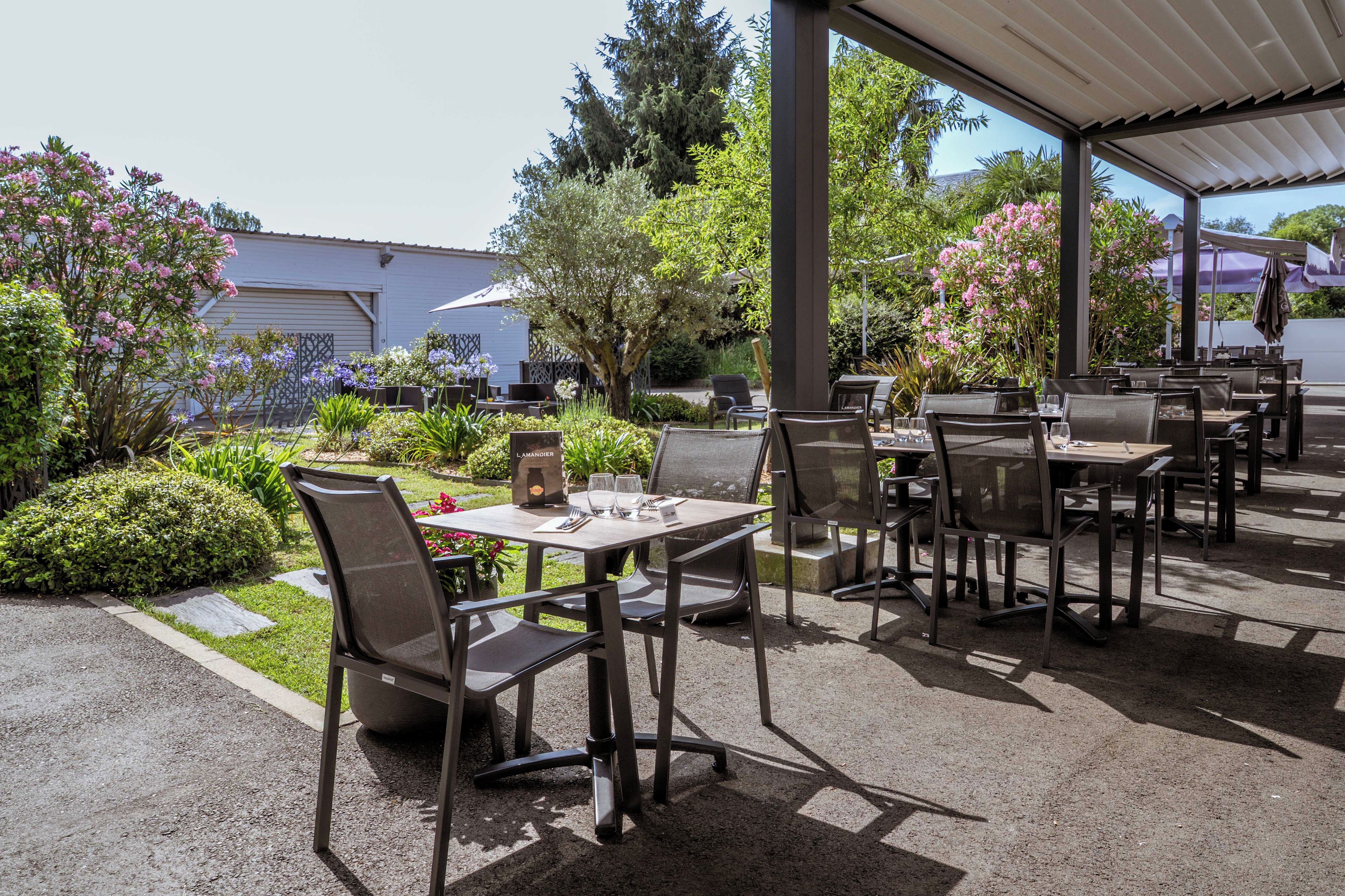 An outdoor patio area with tables and chairs, surrounded by greenery and a building in the background.
