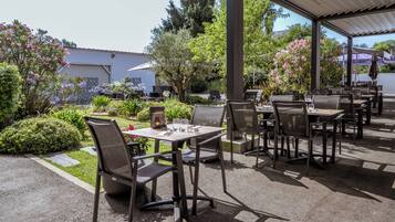 An outdoor patio area with tables and chairs, surrounded by greenery and a building in the background.