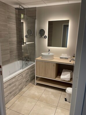 A modern bathroom with a glass-enclosed shower, a freestanding bathtub, a round mirror, and a sink with a wooden vanity. - Hôtel l'Amandier (Morlaas)