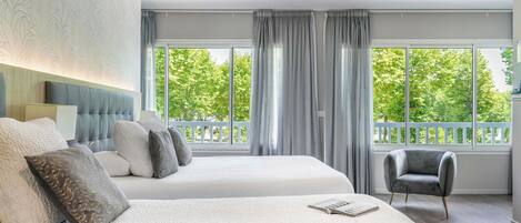 A hotel room with two beds, a grey armchair, and large windows with a view of greenery.