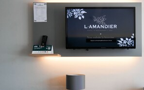 A television screen with the hotel name 'L'AMANDIER' and a welcome message. A remote control and a set of keys are placed on a shelf below the TV. - Hôtel l'Amandier (Morlaas)