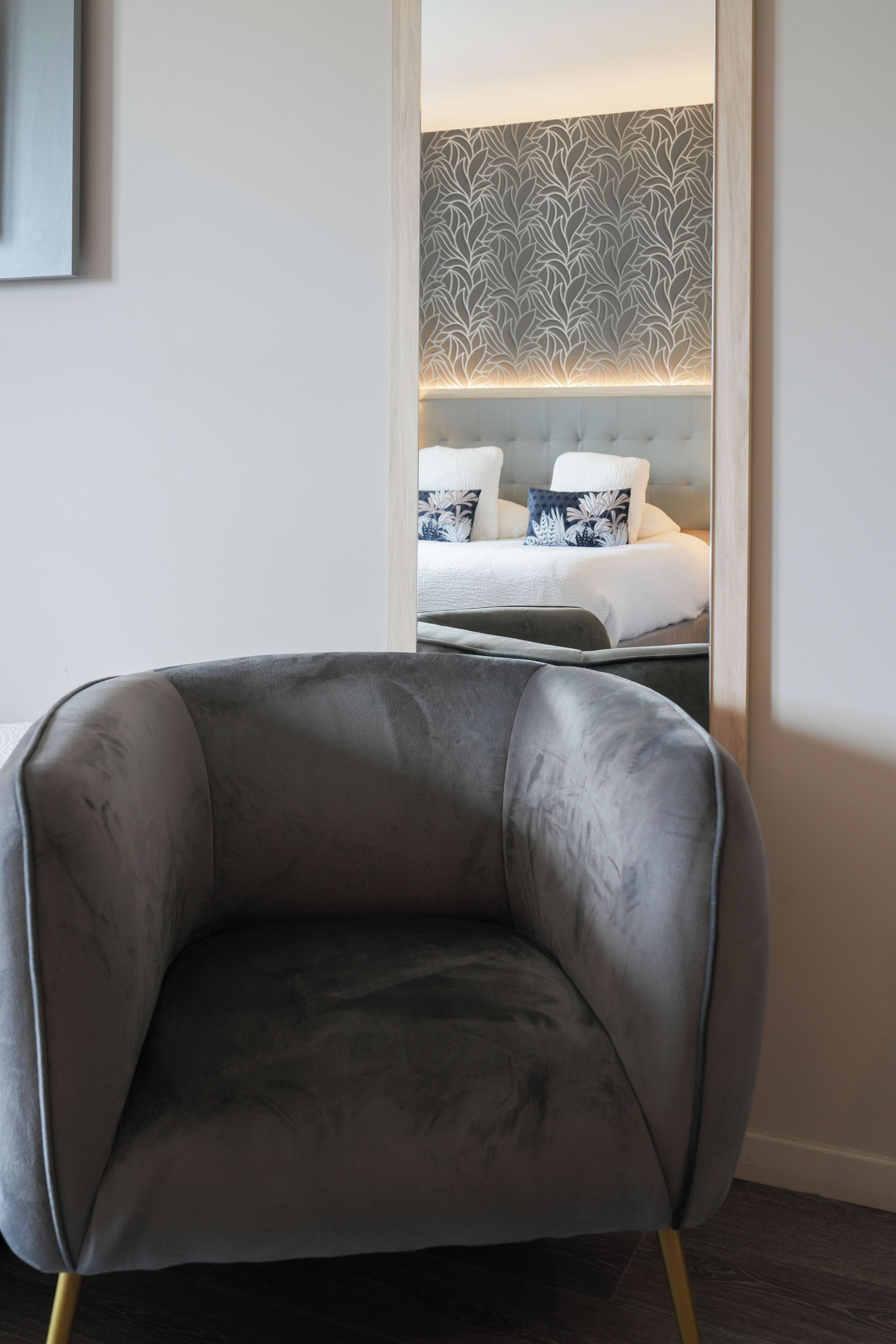 A modern hotel room with a grey upholstered armchair, a bed with white bedding and patterned pillows, and a decorative wall with a leaf-like pattern.