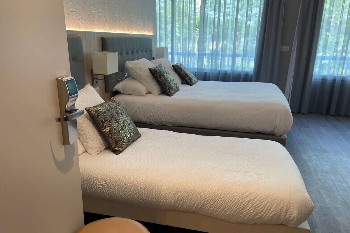 Habitación de hotel con dos camas, una mesita de noche, una lámpara de pared y un ventanal con cortinas.