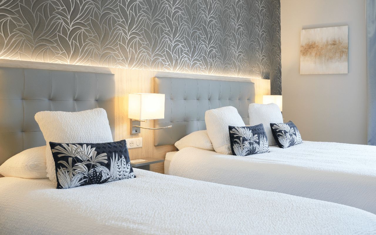 Habitación de hotel con dos camas, cada una con cabecera blanca y almohada estampada con motivos florales.