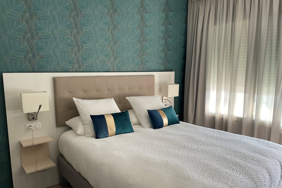 Una cama bien hecha con dos almohadas, una mesita de noche con una lámpara, y una pared con un diseño de hojas.