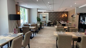 A modern restaurant with multiple dining tables and chairs, a bar area, and a large window providing natural light. - Hôtel l'Amandier (Morlaas)