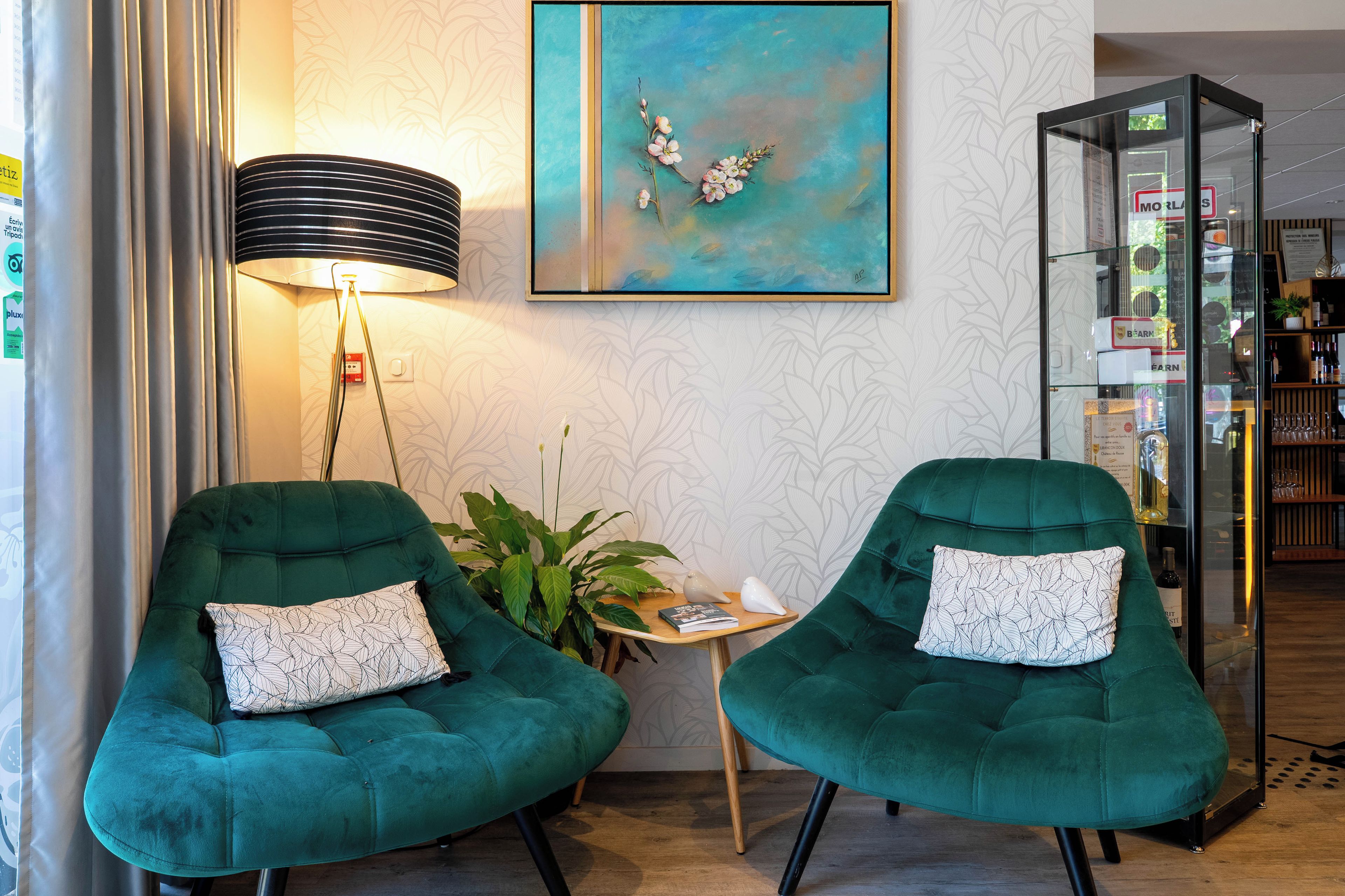 Een kamer met twee groene fauteuils, een tafeltje, een lamp, een plant en een glazen vitrine met spullen.