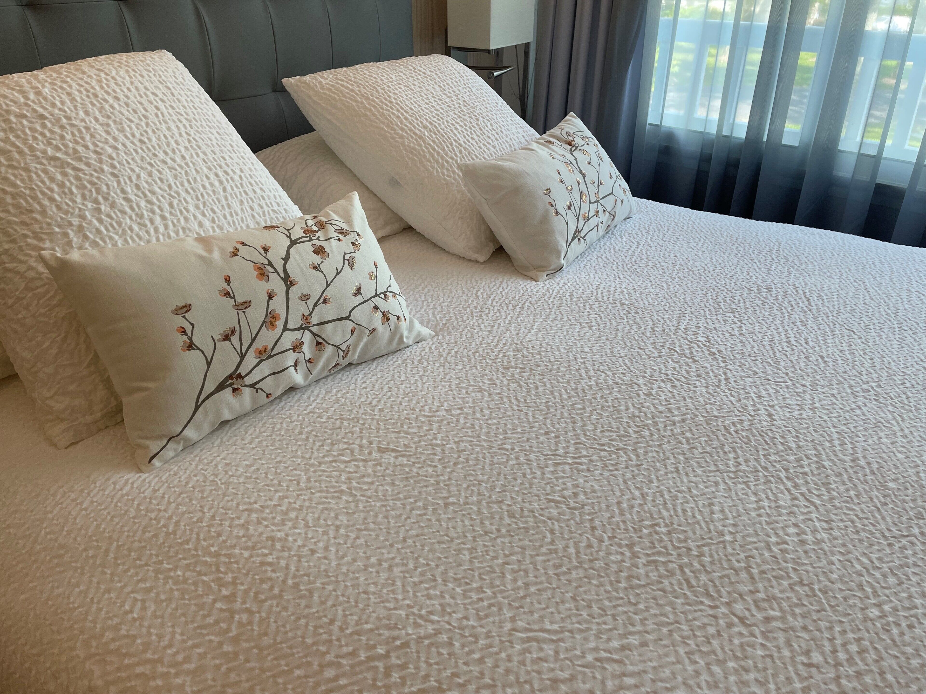 Una cama con una colcha blanca con textura y dos almohadas, una lisa y otra con diseño floral.