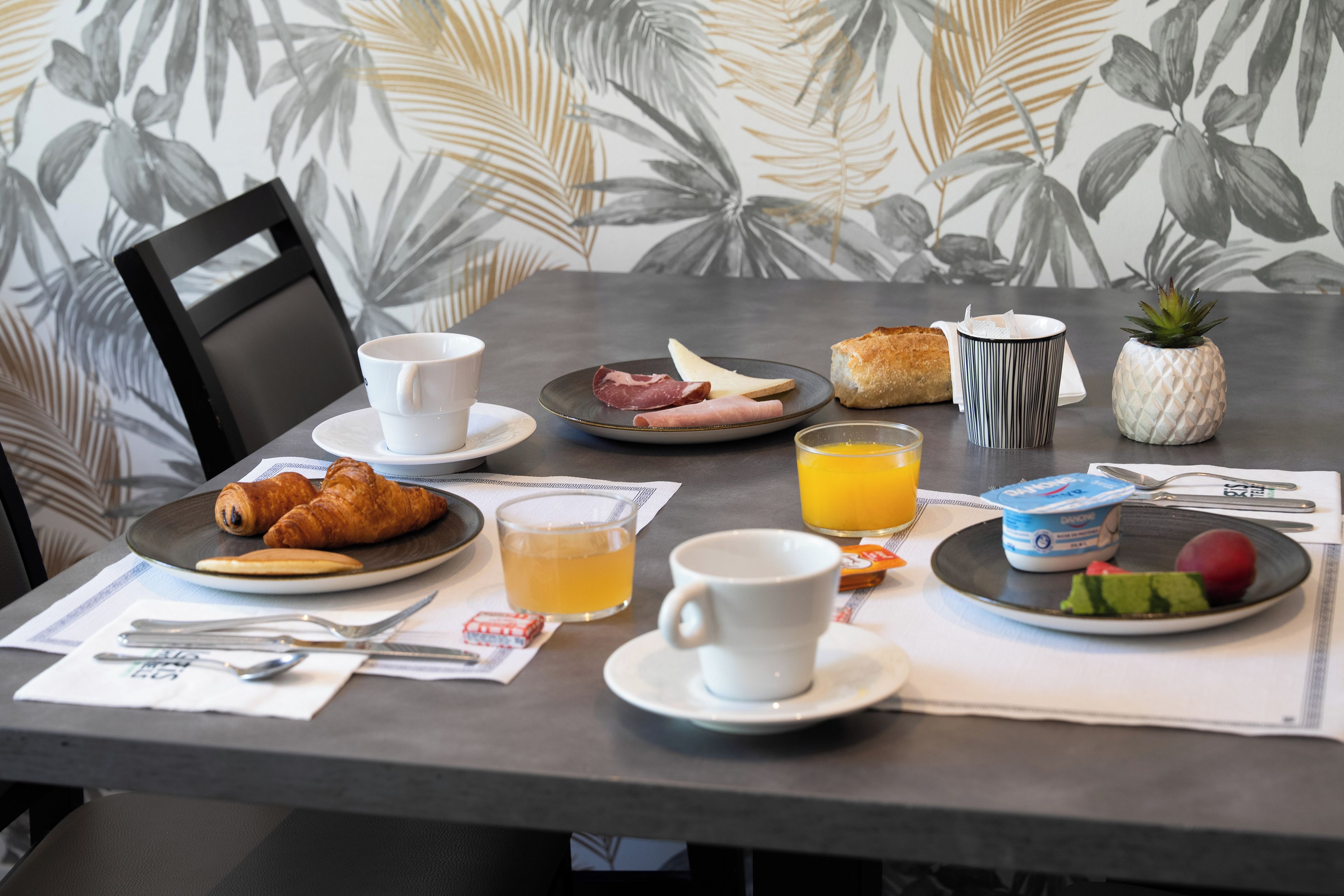 Une table de petit-déjeuner avec des croissants, une assiette de jambon, un verre de jus d’orange, une tasse en forme d’ananas et une petite plante en pot.