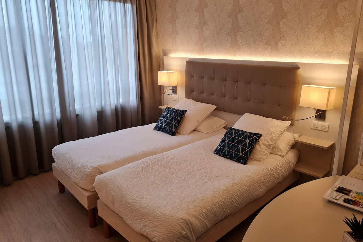 Una habitación de hotel con dos camas, una mesita de noche con lámparas, un revistero y una pequeña mesa con una bandeja.