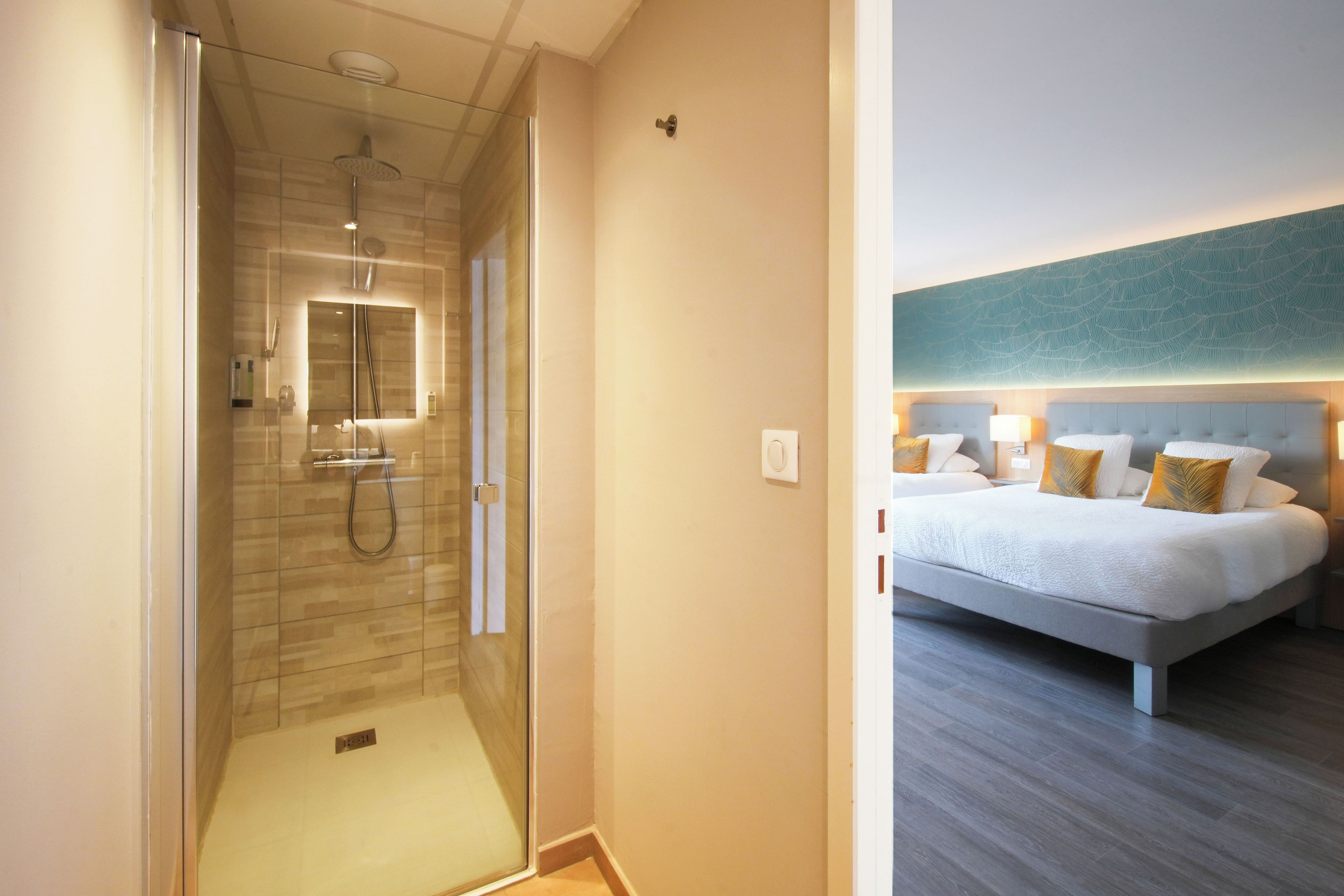 Habitación de hotel con ducha envuelta en vidrio, cama con varias almohadas y lámpara de pared.