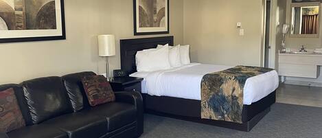 A hotel room with a bed, a black leather sofa, a nightstand with a lamp, and two framed pictures on the wall.