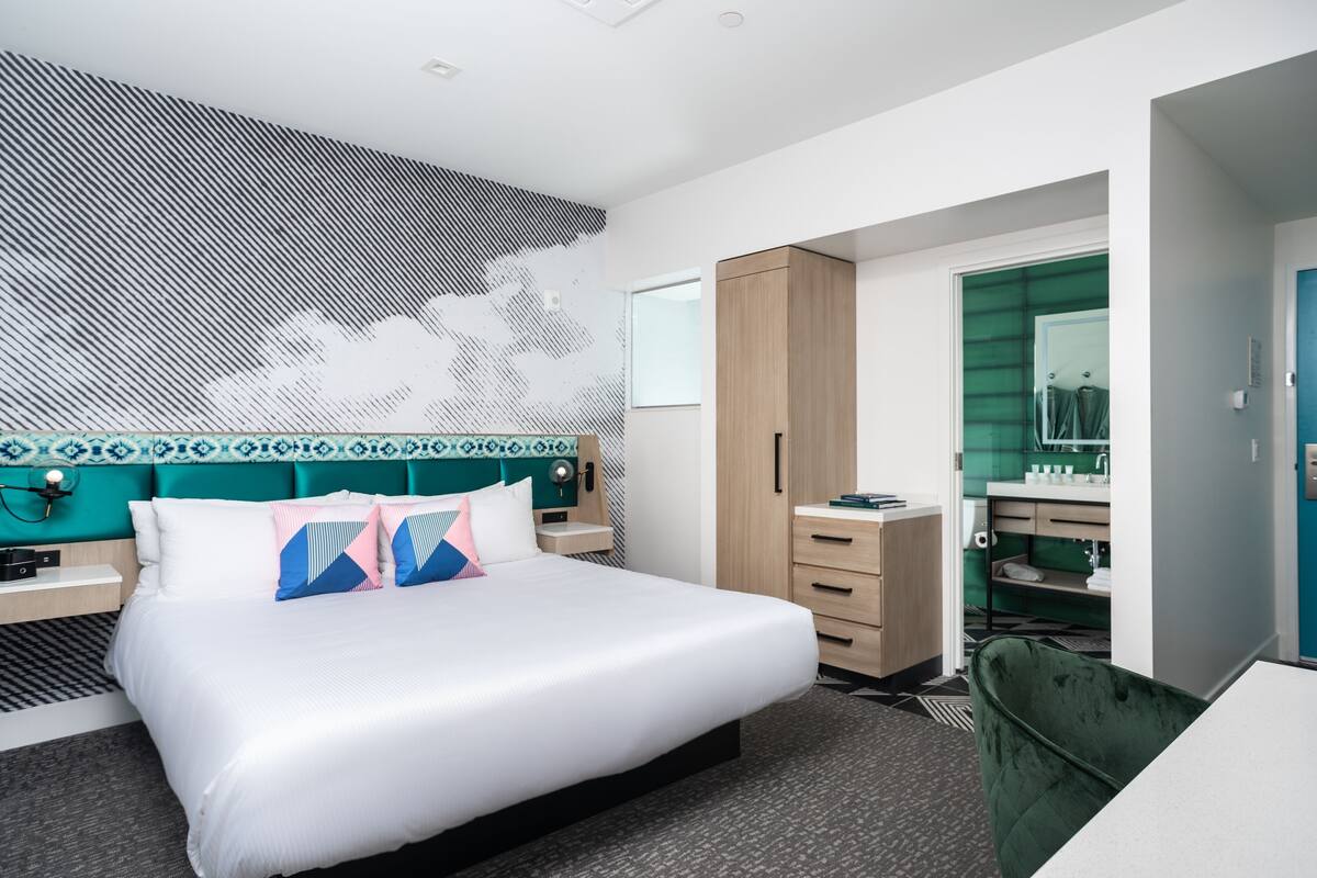 A modern hotel room with a large bed, a wooden wardrobe, a green chair, a desk, and a bathroom with a green sink and mirror.
