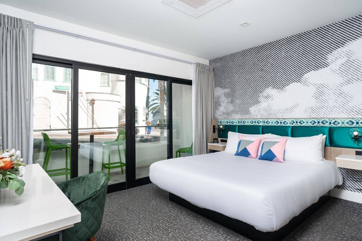 A hotel room with a large bed, a desk with a chair, a glass door leading to an outdoor area, and a patterned wall.