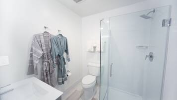 Un baño moderno con ducha envuelta en vidrio, un lavabo blanco y dos batas colgadas en un perchero.