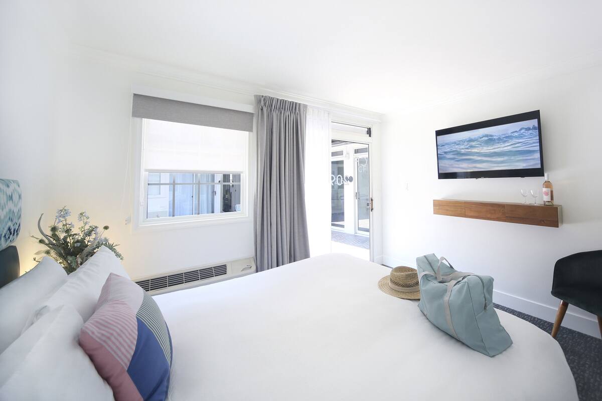 A hotel room with a large bed, a TV mounted on the wall, a small seating area, and a view of the outdoors through a sliding door.
