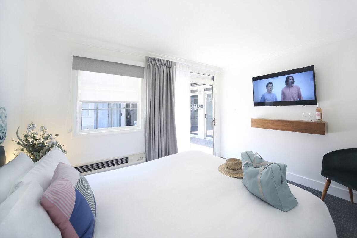 A hotel room with a bed, a television, a small table with a bottle and glasses, a chair, and a window with curtains.