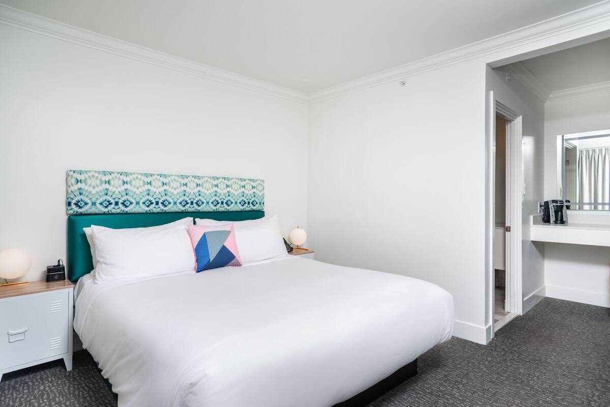 A neatly arranged hotel room with a large bed, a headboard with a geometric pattern, a bedside table with a lamp, and a mirror above the bed.