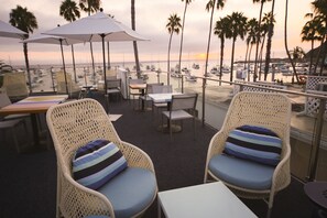 A terrace with wicker chairs and tables, overlooking a marina with boats and palm trees during sunset. - Bellanca Hotel (Avalon)