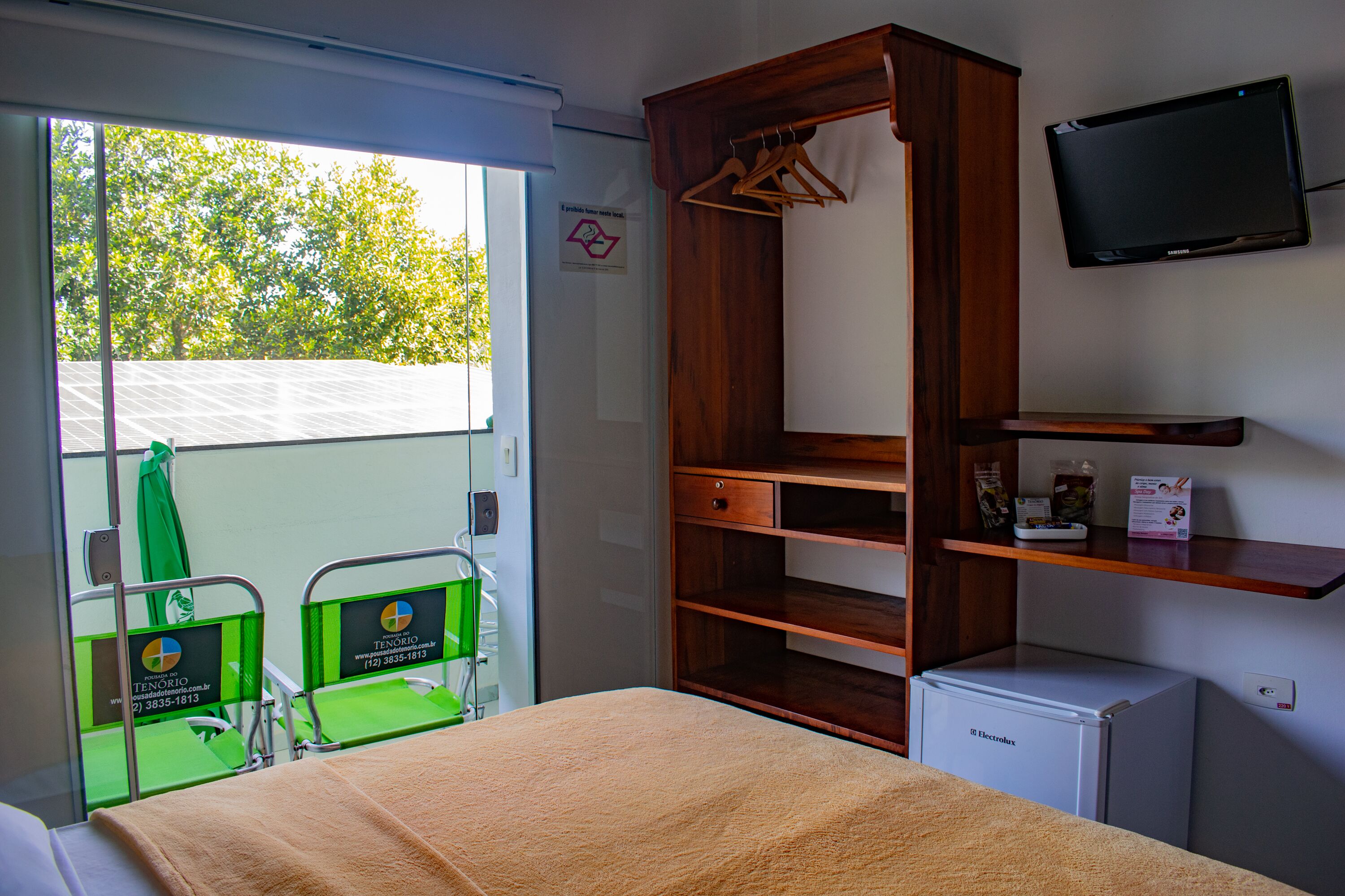 Habitación de hotel con cama, armario de madera, refrigerador pequeño, televisor y ventana con vistas a árboles.