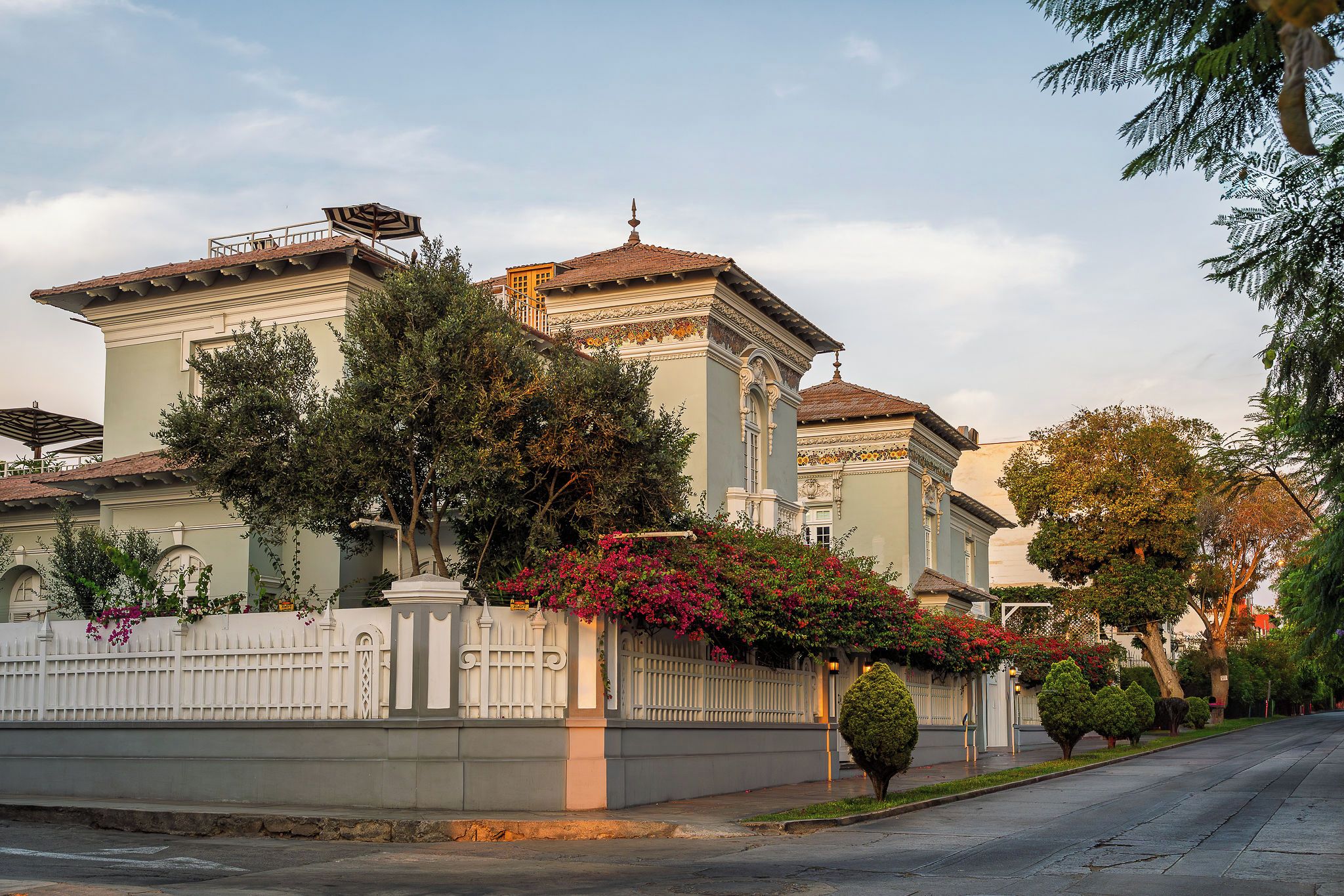 Um edifício de dois andares com cerca branca, varanda e uma árvore com flores cor-de-rosa.
