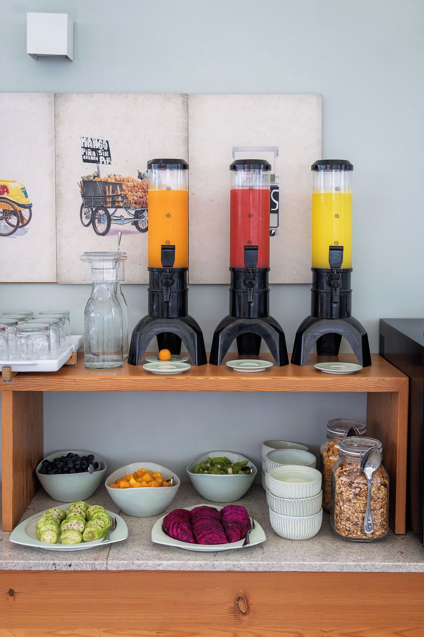 A kitchen counter with four fruit juicers, a glass pitcher, and various food items like fruits and grains.