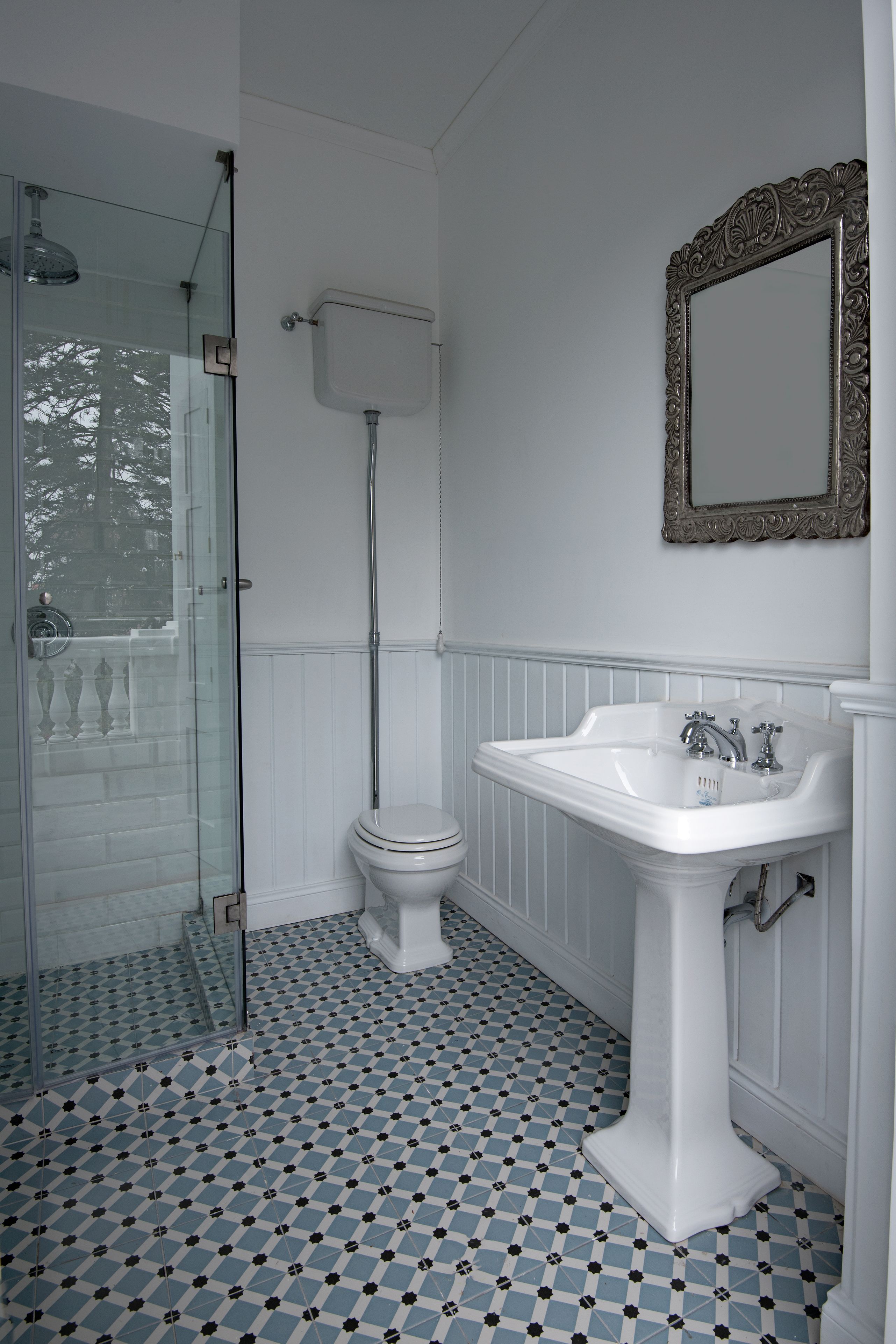 Un baño con cabina de ducha de vidrio, inodoro blanco, lavamanos pedestal y un espejo decorativo.