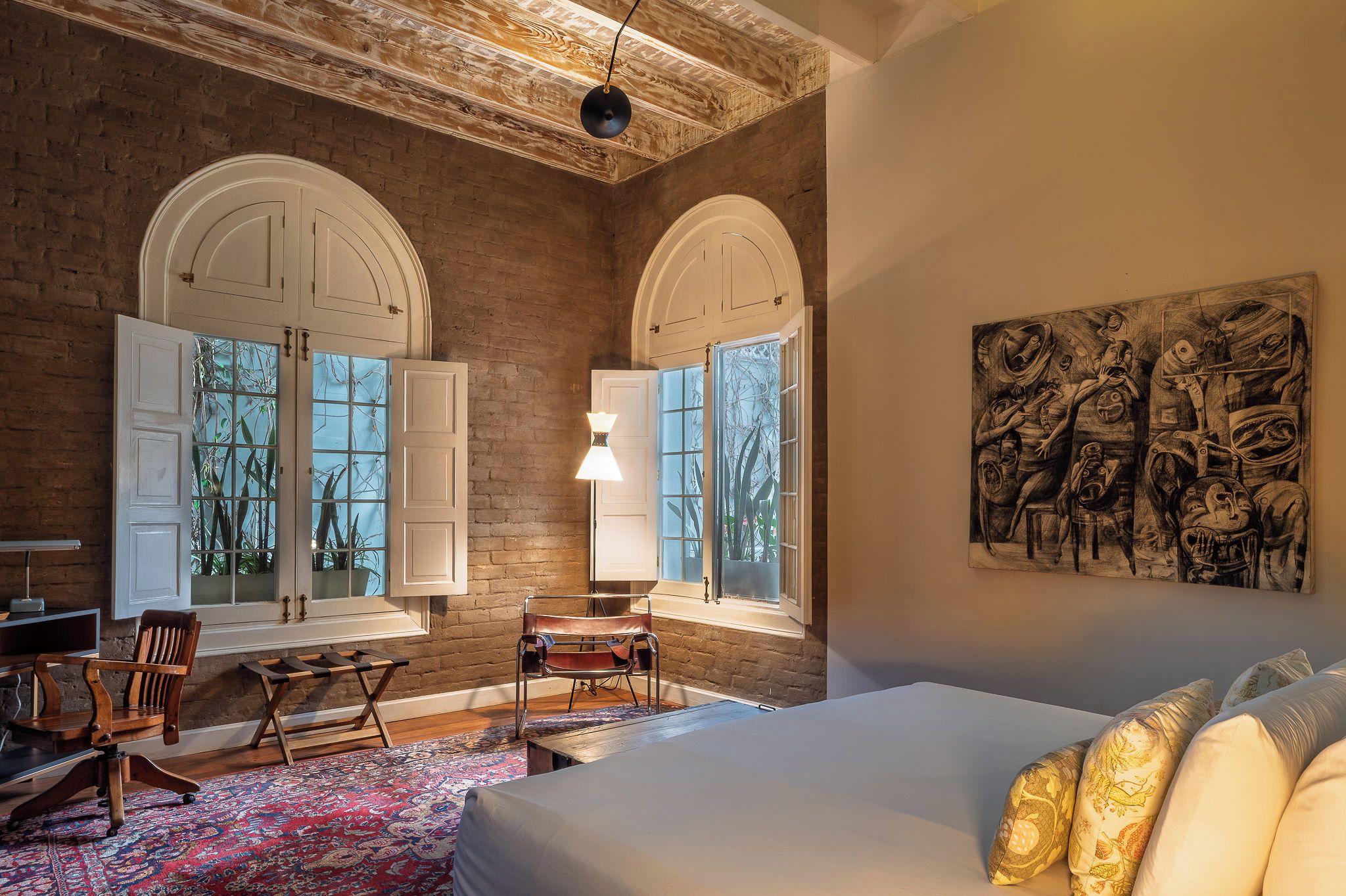 Une chambre avec un grand lit, deux fenêtres avec des volets blancs, un bureau avec une chaise et un tableau accroché au mur.