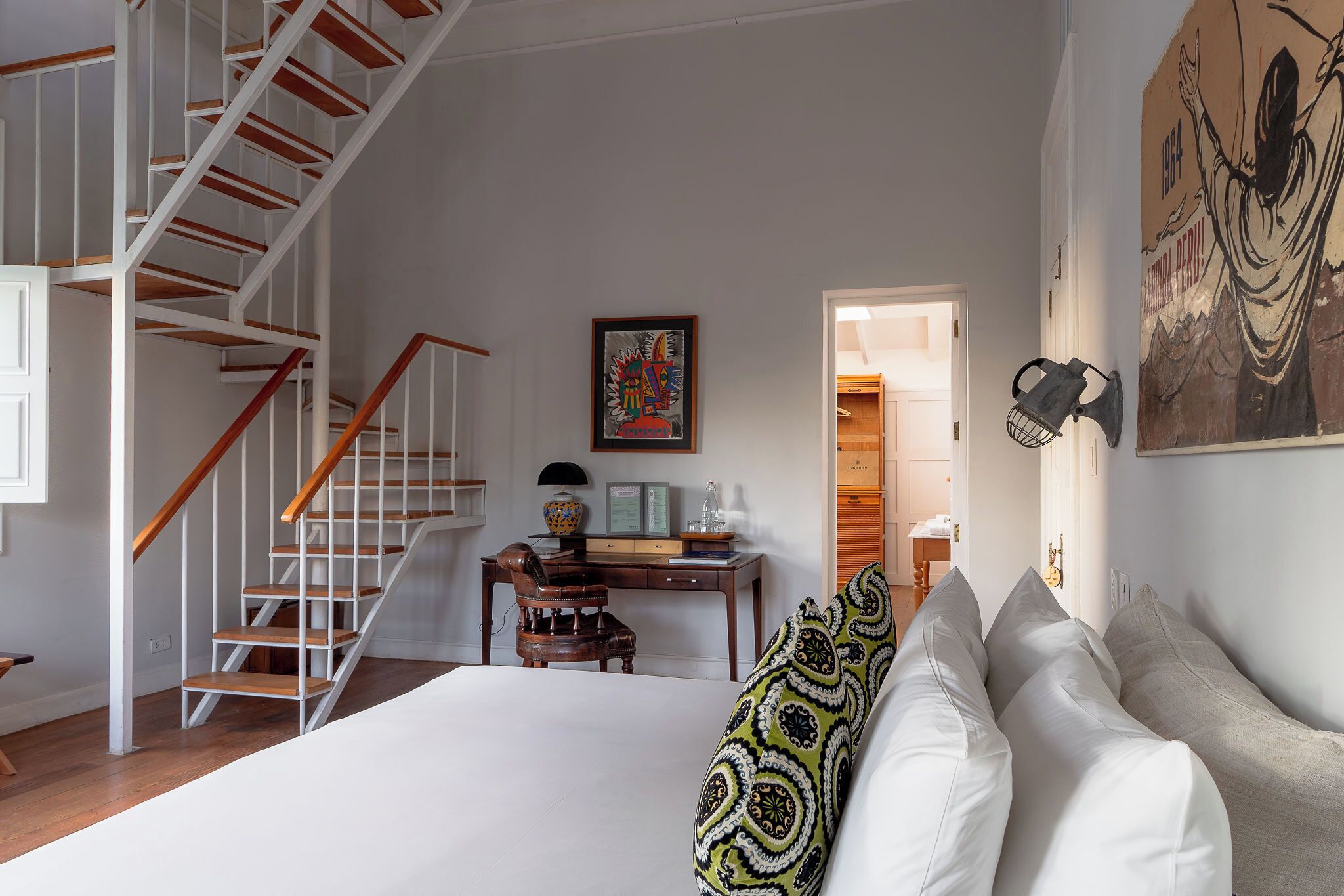 A bedroom with a bed, a sofa, a desk, a chair, a staircase, and a door leading to another room.