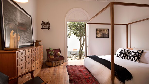Una cama con dosel, un cojín de estampado blanco y negro, un aparador de madera con cajones, una alfombra roja y negra y un cuadro grande en la pared.