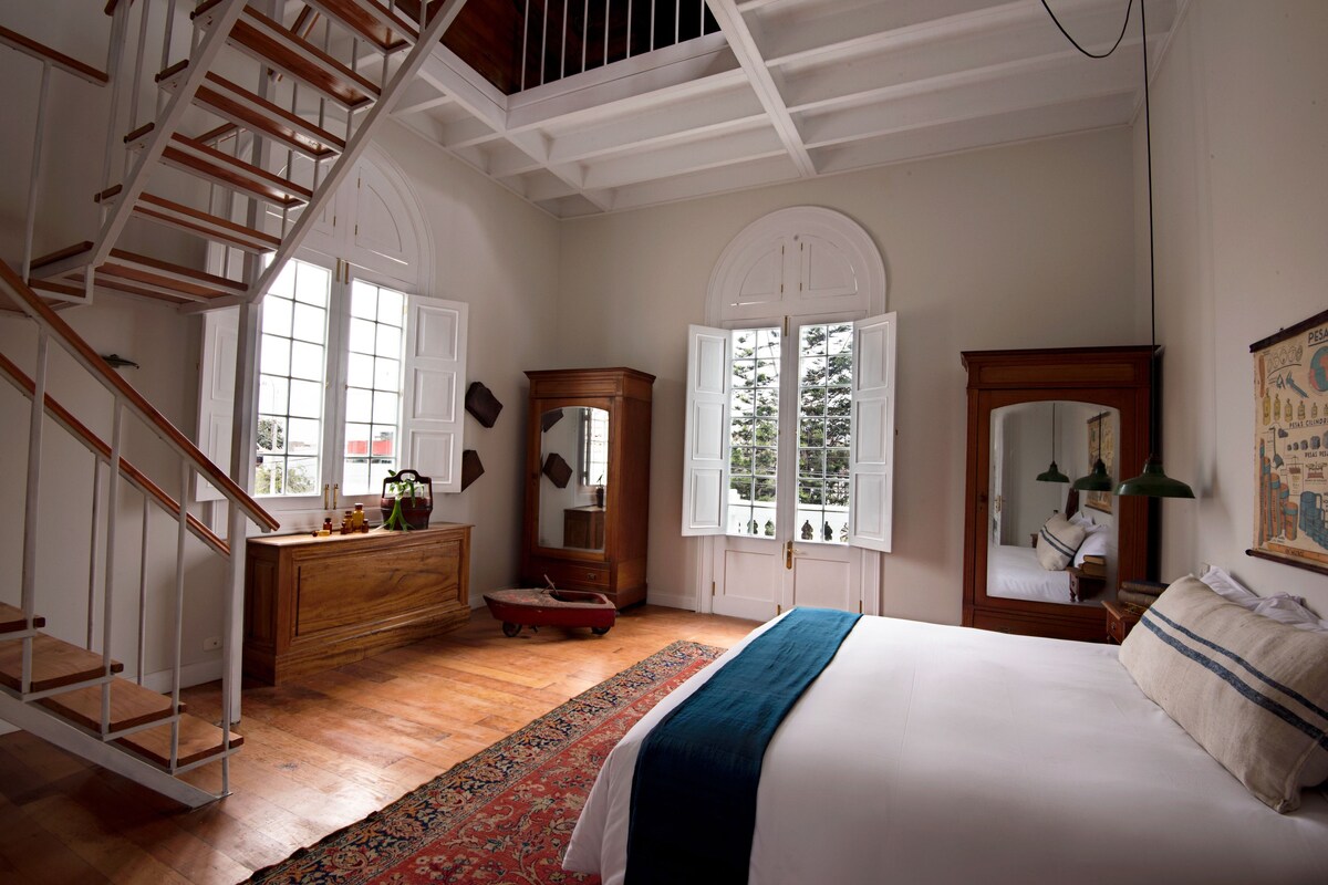 Une chambre avec un escalier en bois, un grand lit, une commode, un miroir et une fenêtre donnant sur des arbres.
