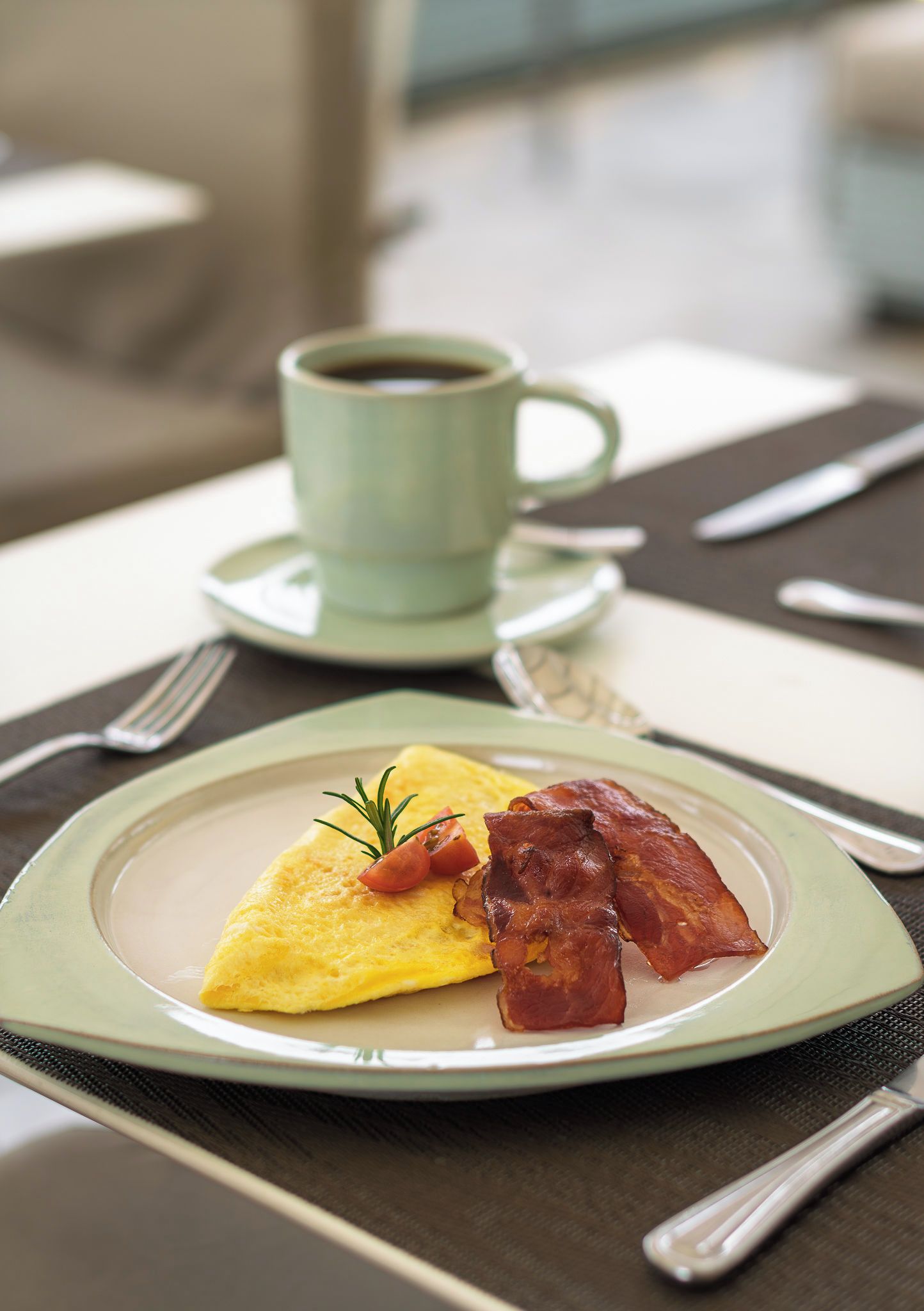 Un plato con tortilla, tocino y una ramita de romero, acompañado por una taza de café sobre una mesa.